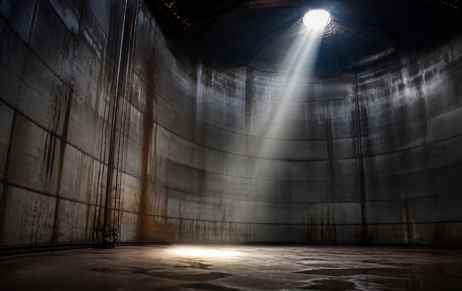Interior of a large empty steel storage tank lit by a single shaft of light through the roof manway