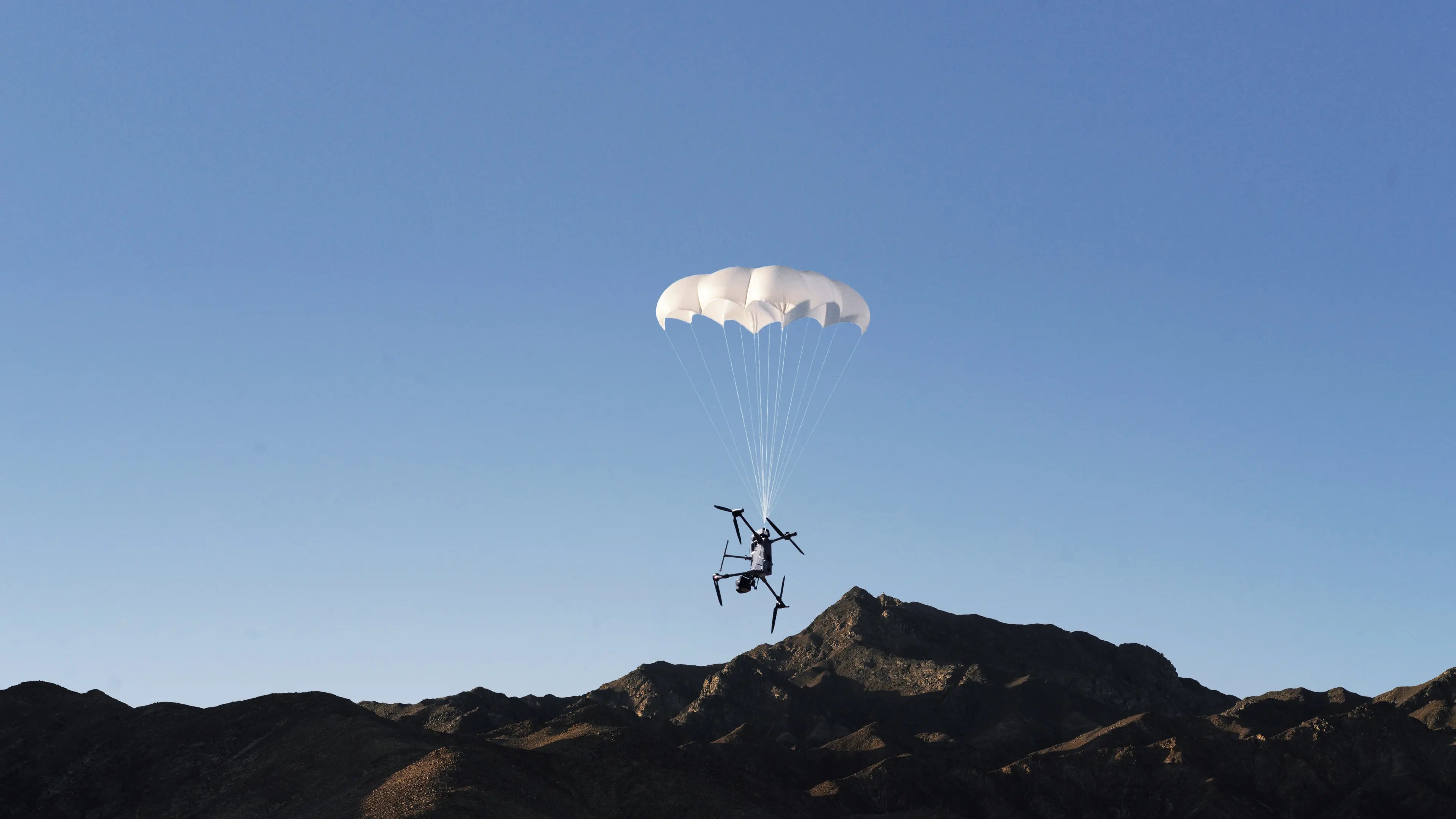 DJI Matrice 400 descending under a deployed DJI AP100 Parachute canopy over mountain terrain.